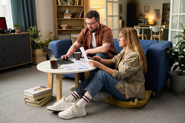 Caucasian young adult man and Caucasian young adult woman sitting on floor in living room reviewing...