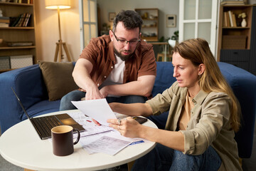 Caucasian middle aged man and Caucasian middle aged woman reviewing tax documents together at table, man pointing at paper while woman holding paperwork, laptop and calculator visible