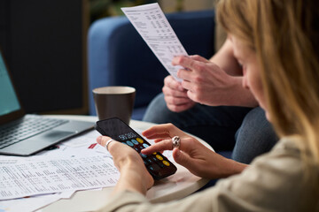 Caucasian young woman using smartphone calculator while reviewing tax documents with Caucasian man, both sitting at table with financial papers and laptop, focusing on paying taxes