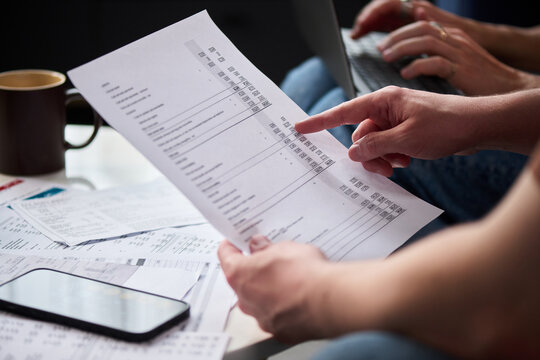 Caucasian middle aged man reviewing tax documents and using laptop while sitting at desk, holding printed financial statement, surrounded by paperwork and smartphone, focusing on paying taxes