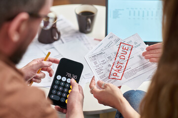 Middle aged Caucasian man and middle aged Caucasian woman reviewing overdue tax bills, holding smartphone with calculator app and past due notice, sitting at table with paperwork