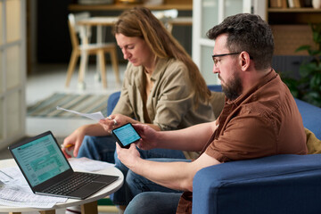 Middle aged Caucasian man using smartphone and laptop while reviewing tax documents with young adult Caucasian woman sitting beside him, both focusing on paying taxes at home