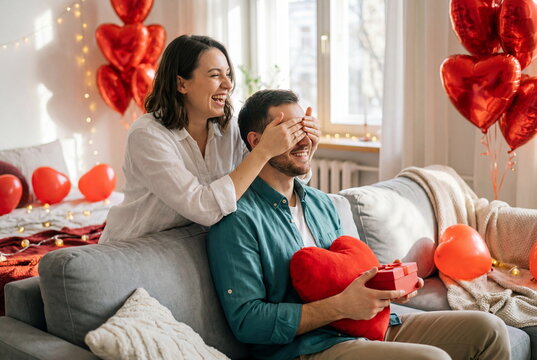 Happy woman giving a surprise gift to her smiling boyfriend, covering his eyes on Valentine's Day. Romantic love and anniversary celebration at home.