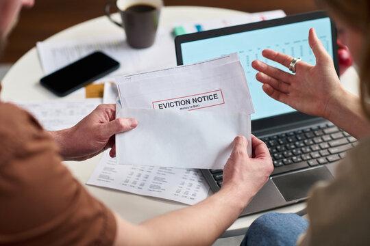 Caucasian young adult man holding eviction notice envelope while sitting at desk with financial documents and laptop, Caucasian woman gesturing with hand discussing paying taxes - Powered by Adobe
