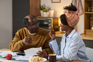 Middle aged Black man discussing financial documents with young adult Black woman at kitchen table, both reviewing tax forms and using calculator, paperwork and bills visible