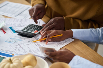 Closeup of Black man and Black woman calculating taxes together using calculator and reviewing financial documents at table, hands holding pencil and receipts, paperwork scattered around