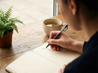 Young woman writing in gratitude journal at cafe mental health self reflection therapy planning diary notebook pen mindfulness lifestyle positive thinking handwriting personal growth