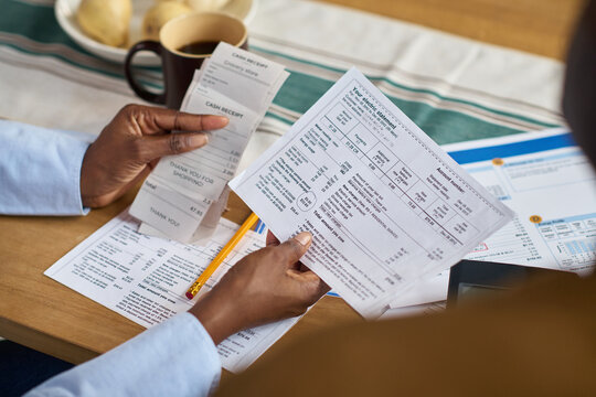 Middle aged Black woman holding receipts and tax documents while sitting at table, reviewing financial paperwork and calculating expenses for paying taxes, partial hands visible