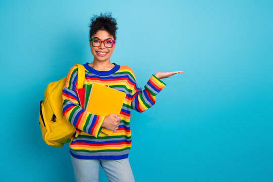 Young girl student with colorful striped sweater and yellow backpack smiles blue background presenting notebooks for education and fashion lifestyle