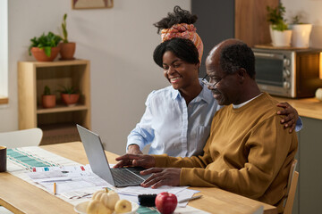 Middle aged Black man and young adult Black woman sitting at table using laptop together, smiling while reviewing financial documents and tax forms, working on paying taxes at home