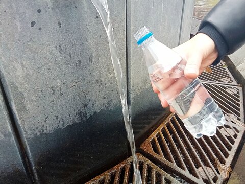 A person filling a clear plastic bottle with fresh running water from an outdoor tap. The scene captures everyday access to clean water, hydration and practical outdoor use. Suitable for themes of sus