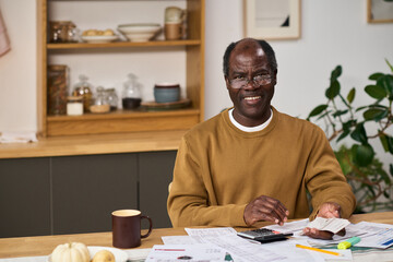 Middle aged Black man sitting at table reviewing financial documents and using calculator while...