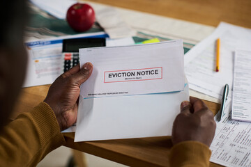Middle aged Black man opening eviction notice letter at desk, surrounded by financial documents and...