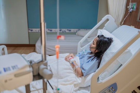 Sick Asian girl wearing oxygen mask lying on hospital bed receiving treatment. Healthcare concept, respiratory disease, hospital admission, medical care, and child patient in emergency situation. - Powered by Adobe