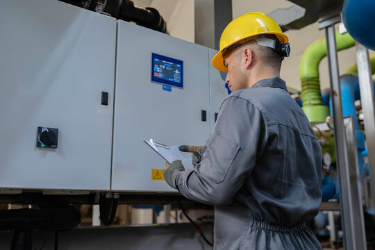 Professional technician inspecting electrical control panel in a factory plant. Male engineer recording operational data on clipboard during safety maintenance of industrial machinery system.