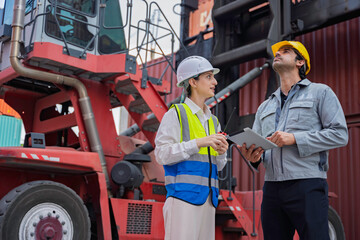 Two logistics engineers discussing work plan at container terminal. Female supervisor pointing up while male colleague checks data on digital tablet during cargo inspection at shipping port.
