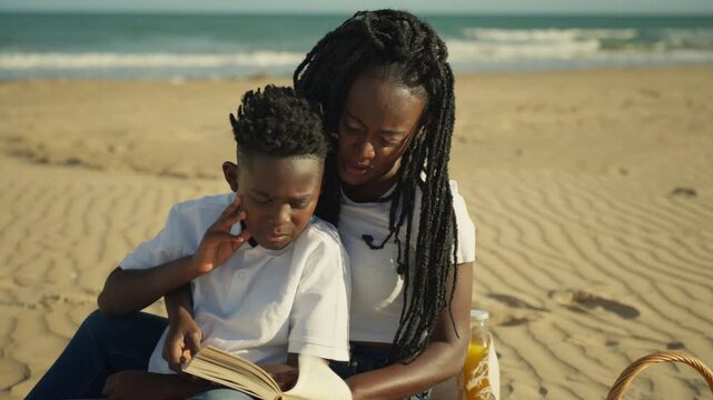 Young african american mother and son sit on sandy beach reading and picnicking, sharing peaceful bonding moment of learning, laughter and summer relaxation by sea