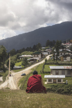 Gangtey Monastery, Bhutan - 21 September 2025: View of a lone monk in vibrant red robes contemplates the Phobjikha Valley's village nestled amidst rolling green hills and cloudy skies.