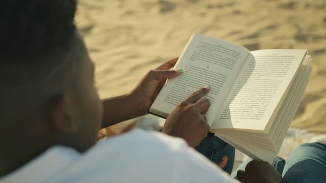 Over the shoulder view of person read book while sitting on sand, pointing at text with a finger. Relaxing hobby on sunny day, focusing on literature and self education outdoors