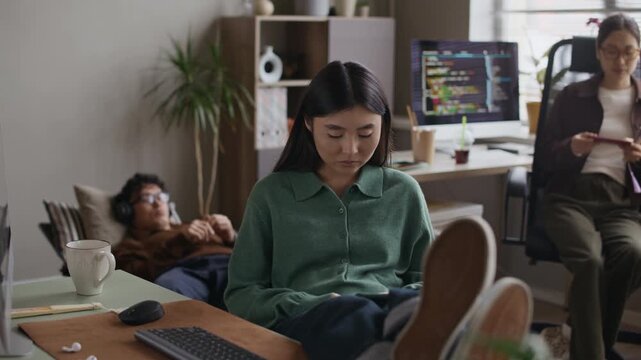 Handheld shot of young relaxed Asian woman using mobile phone at working table while Asian colleagues resting in background at startup office space