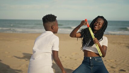 Joyful african american mother and young son having fun together on sandy beach, with woman blowing large soap bubbles and laughing boy trying to catch them during sunny day - Powered by Adobe
