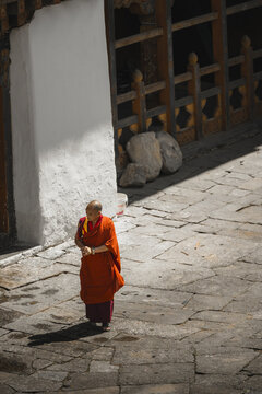 Trongsa, Bhutan - 22 September 2025: View of a monk in vibrant saffron robes walking across the textured stone courtyard, framed by the stark white walls and dark shadows.