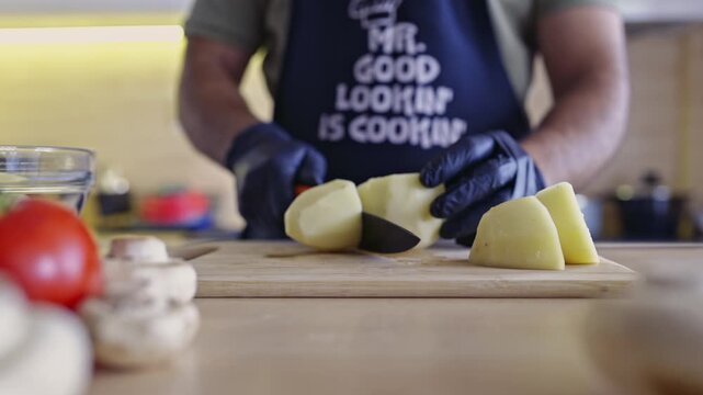 A man wearing an apron and black gloves, precisely cuts peeled potatoes on a wooden board. The clear sounds of the knife on the board add an authentic culinary touch to this preparation scene.