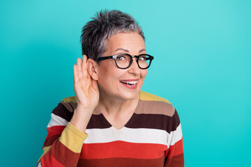 Happy mature woman with short gray hair and glasses listening with hand to ear against a blue...