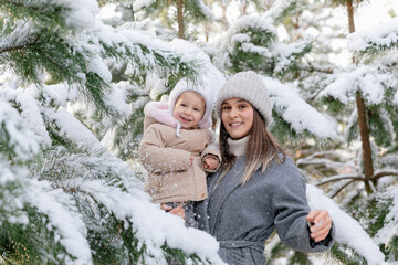 Obraz premium Mother holding her laughing toddler in a snowy pine forest. Joyful winter moment as snow falls from the branches.