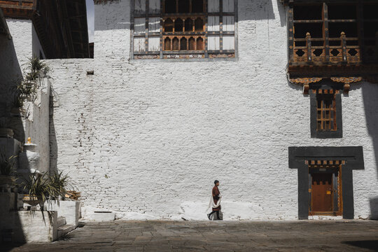 Fototapeta Trongsa, Bhutan - 22 September 2025: View of whitewashed walls contrasting with the intricate dark wooden window frames and a solitary figure walking past the traditional architecture.