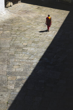 View of a lone monk clad in vibrant saffron robes walks across the stone courtyard, half bathed in sunlight, half shrouded in shadow, Trongsa, Bhutan.