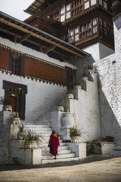 View of a lone monk ascends worn stone steps alongside whitewashed walls and traditional Bhutanese architecture, a vibrant splash of red against the muted tones, Trongsa, Bhutan.