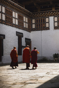 Trongsa, Bhutan - 22 September 2025: View of three monks in vibrant red robes traversing the sun-drenched stone courtyard, framed by the monastery's white walls and intricate wooden architecture.