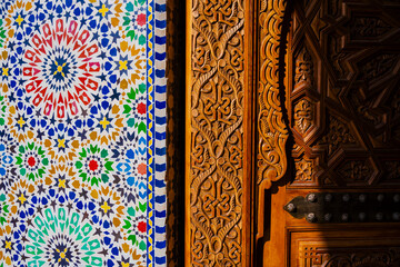 Intricate mosaic tiles beside ornate wooden carvings in vibrant sunlight. Marrakesh, Morocco