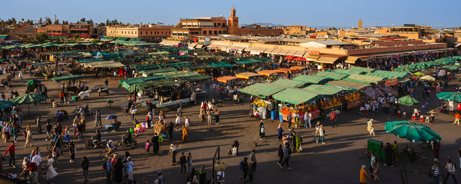Bustling outdoor market with green stalls, diverse crowd, and historic architecture in the background. Marrakesh, Morocco