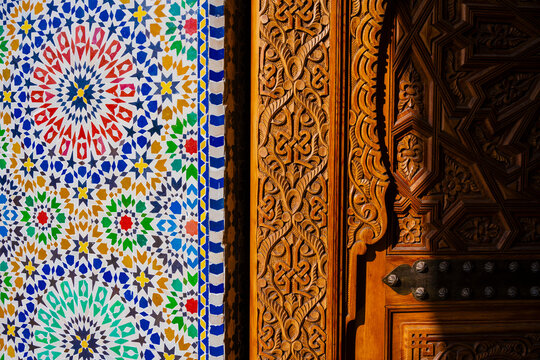 Intricate mosaic tiles beside ornate wooden carvings in vibrant sunlight. Marrakesh, Morocco