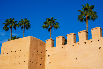 Earthen wall with crenellations and palm trees set against a clear blue sky. Marrakesh, Morocco