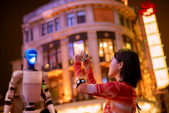 Woman taking photo of humanoid robot at night in city with colorful neon lights. Shanghai, China