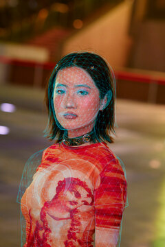 Woman in vibrant attire with projected geometric pattern on face stands in urban setting. Shanghai, China