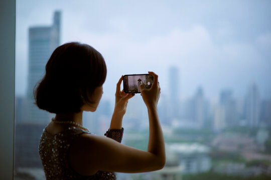 A woman photographs a city skyline through a window on a cloudy day. Shanghai, China