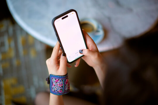 Person with colorful bracelet holding a smartphone with a blank white screen above a table with coffee. Shanghai, China