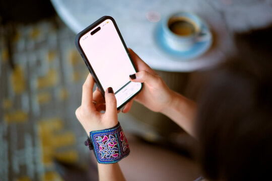 Woman with patterned bracelet holding a smartphone near a table with a cup of coffee. Shanghai, China