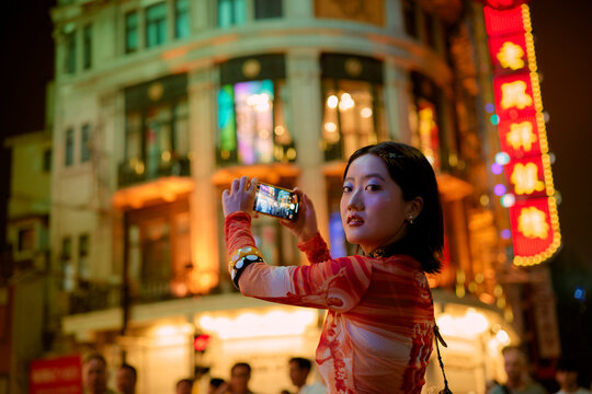 Woman takes a nighttime photo on a vibrant street with neon signs and a large building. Shanghai, China