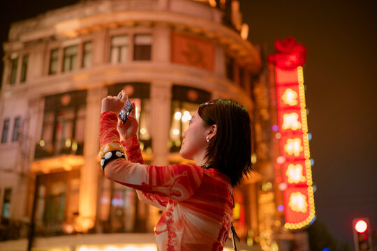 Woman taking photos of brightly lit buildings at night in an urban setting. Shanghai, China
