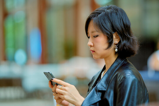 Woman in leather jacket engrossed in smartphone against blurred background. Shanghai, China