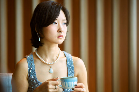 Woman in a blue dress holding a cup, seated indoors with striped background Shanghai, China