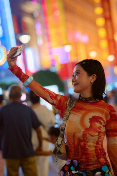 Young woman takes selfie in vibrant, colorful city street at night, smiling brightly. Shanghai, China