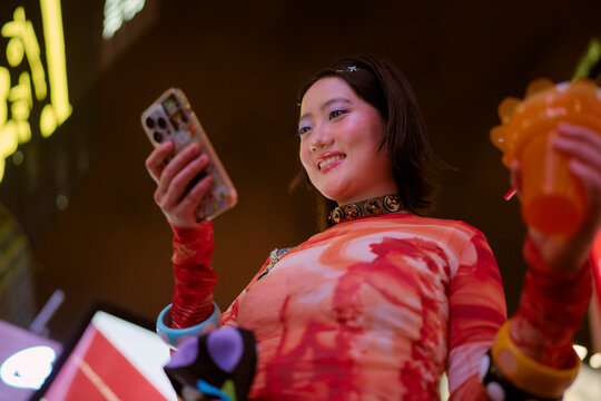 Young adult in colorful outfit smiles at phone while holding a bright drink in a lively setting. Shanghai, China