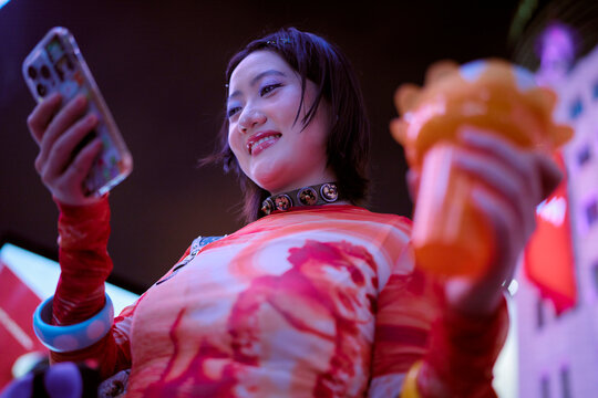 Young woman smiling, holding a smartphone and colorful drink in light-up surroundings. Shanghai, China