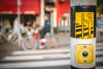 Blurred cyclists and pedestrians at a crosswalk with a focus on a yellow pedestrian button. Amsterdam,. Netherlands